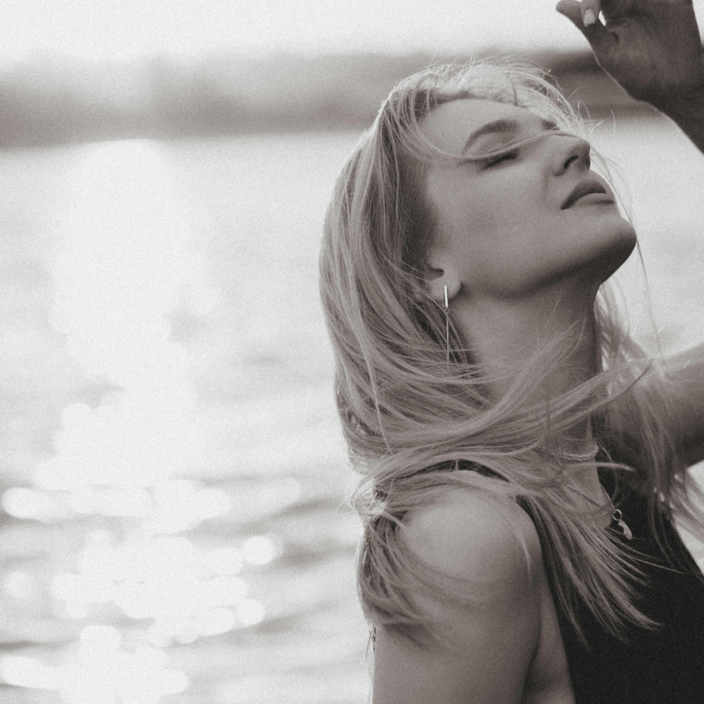 Femme aux yeux fermés profitant d'une douce brise au bord de l'eau, sur une photographie sépia, son diaphragme bougeant doucement à chaque respiration.