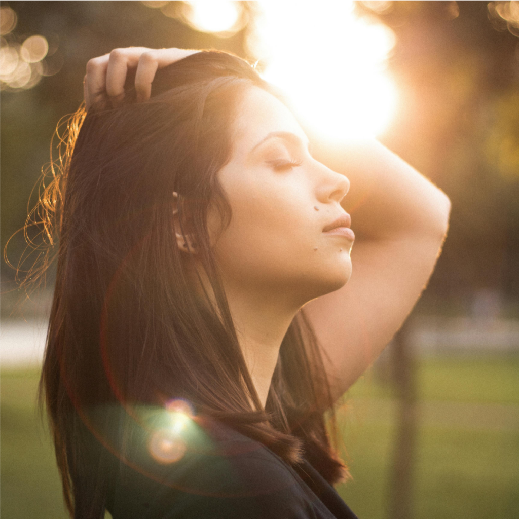 Une femme aux yeux fermés, passant ses doigts dans ses cheveux, baignée dans la chaude lueur du coucher de soleil.