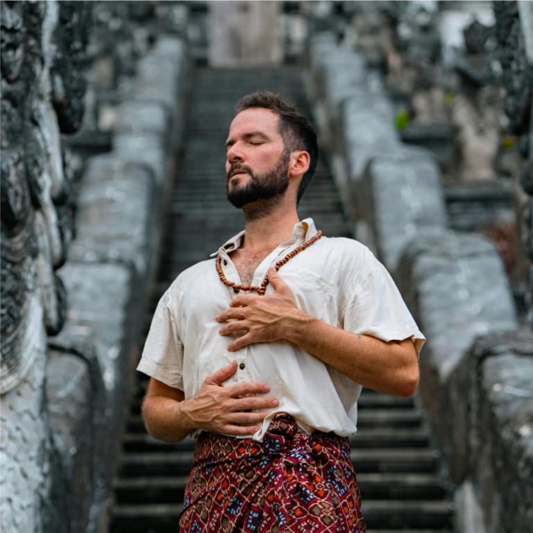 Un homme vêtu d'une chemise blanche et d'un pantalon à motifs, debout, les yeux fermés et la main sur son diaphragme, manifestant un sentiment de calme, avec des sculptures en pierre bordant l'escalier derrière lui.