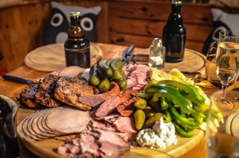 Un plateau en bois avec un assortiment de viandes tranchées, de saucisses, de fromage, de cornichons et de poivrons verts est posé sur une table dressée avec des boissons, des assiettes et des couverts.