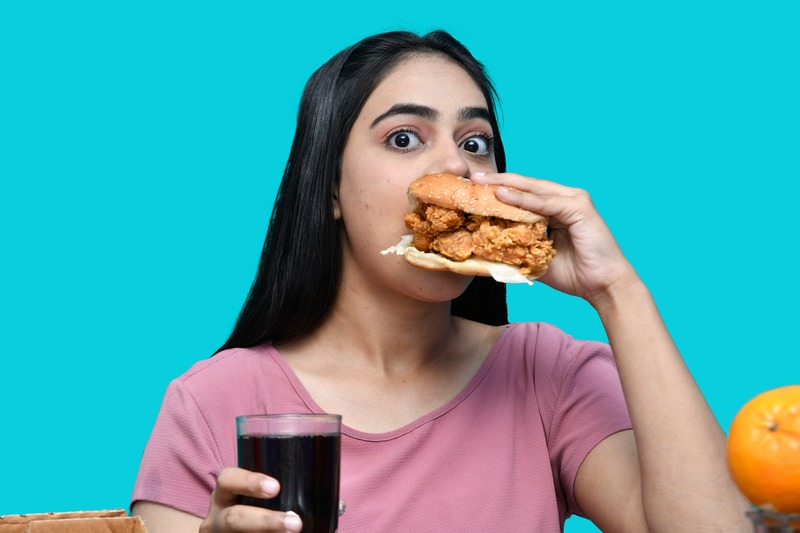Une femme en chemise rose tient un sandwich au poulet frit, symbole de la malbouffe, et un verre de boisson foncée, avec une expression surprise, sur un fond turquoise, faisant allusion aux risques du diabète.