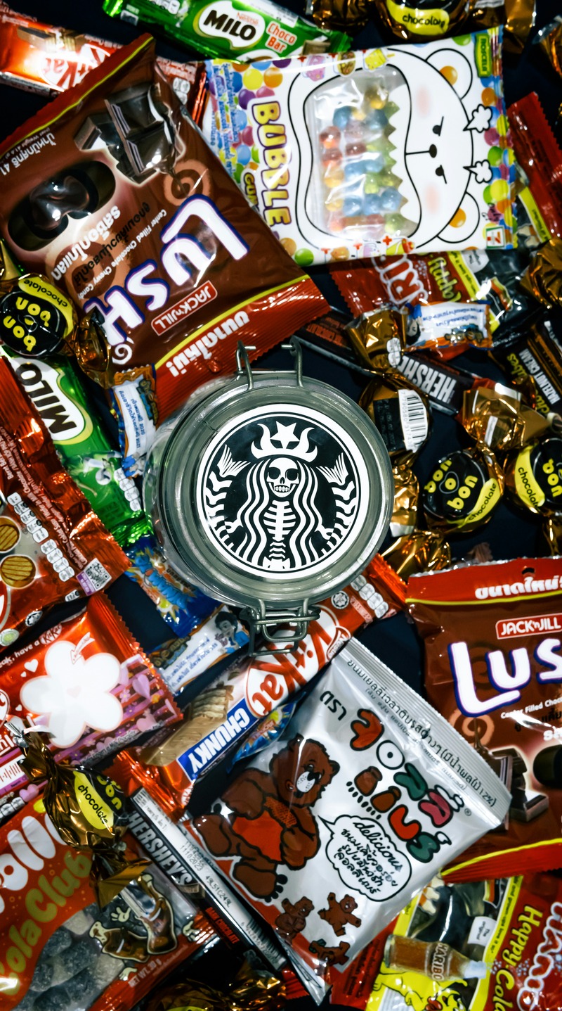 Un assortiment d'emballages de bonbons et de chocolats colorés entoure une boîte en métal avec un logo noir et blanc ressemblant à la sirène de Starbucks - une vue tentante, mais à apprécier avec modération pour ceux qui font attention au diabète.